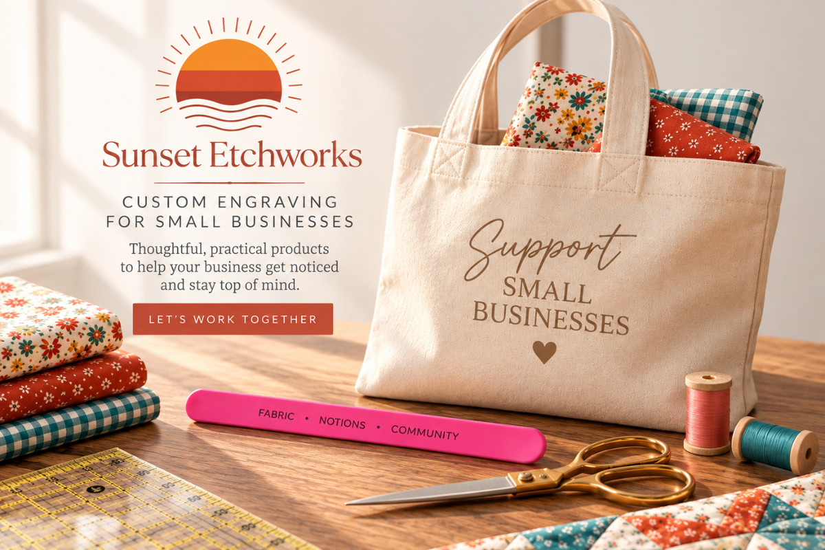 A beige canvas tote bag with custom gold engraving reading 'Support Small Businesses' and a small heart, styled on a wooden table with sewing supplies. The tote bag contains patterned fabrics.