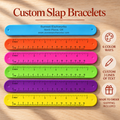 Six colorful plastic ruler-style slap bracelets are shown on a light wood background, with the title 'Custom Slap Bracelets' at the top. The bracelets come in blue, orange, pink, green, purple, and yellow, each printed with measurement markings and sample text.