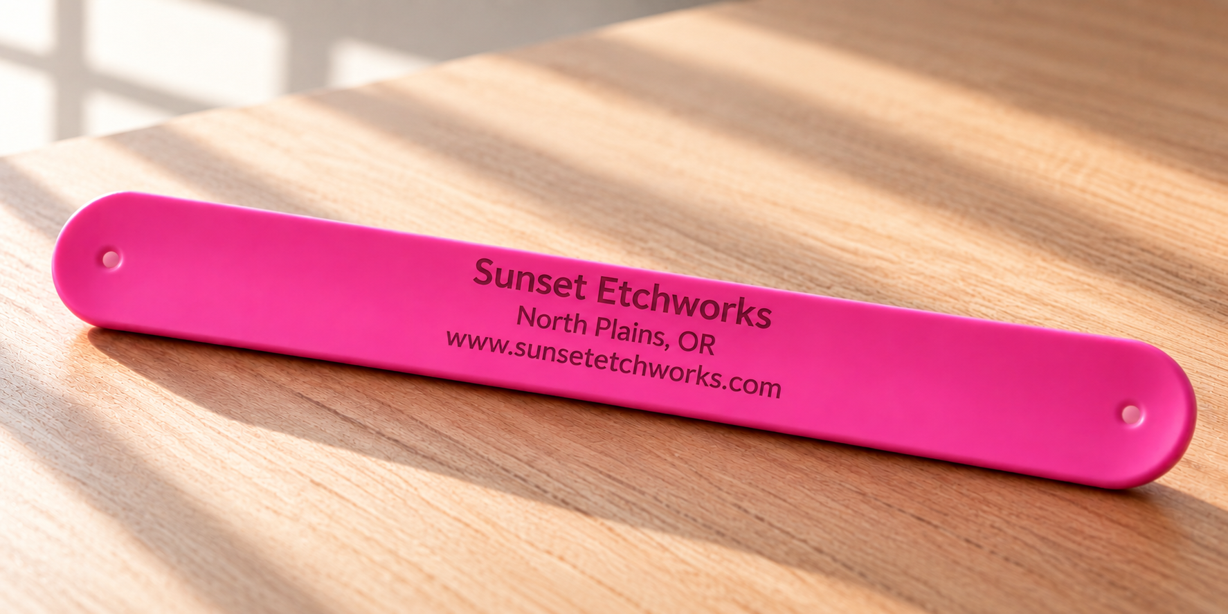 A bright pink rectangular metal tag with rounded corners lies on a wooden tabletop. The tag is printed with the text 'Sunset Etchworks North Plains, OR www.